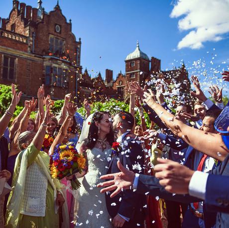 A bride and groom outside sharing a kiss, with a crowd scattering confetti over them. Aston Hall is in the background