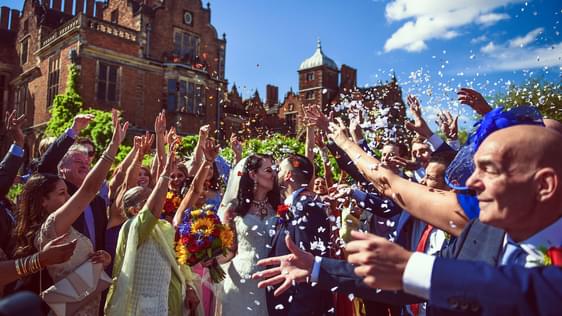 A bride and groom outside sharing a kiss, with a crowd scattering confetti over them. Aston Hall is in the background