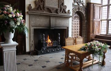 A grand fireplace with a burning fire, in front of which is a table and two chairs. A bouquet of flowers is on the table and more flowers are on a nearby stand.