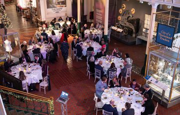 Guests dressed formally sit at circular tables and enjoy a festive dinner event in a decorated, historic venue.