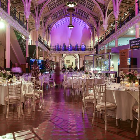 Elegant gallery set up with round tables, white chairs, floral centrepieces, and purple ambient lighting.