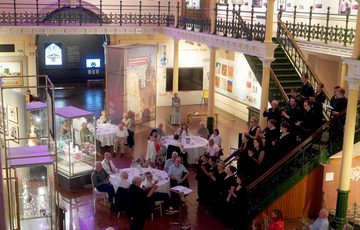 The Industrial Gallery at night set up for an event with people seated at round tables and a choir performing on the stairs.