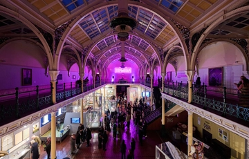 Lots of visitors in two level gallery which has an ironwork ceiling and balcony. The gallery is lit up in purple neon colours.