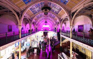 Lots of visitors in two level gallery which has an ironwork ceiling and balcony. The gallery is lit up in pink neon colours.