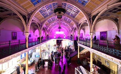 Lots of visitors in two level gallery which has an ironwork ceiling and balcony. The gallery is lit up in pink neon colours.