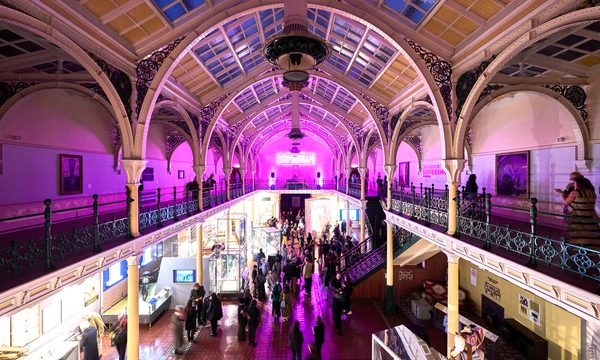 Lots of visitors in two level gallery which has an ironwork ceiling and balcony. The gallery is lit up in pink neon colours.