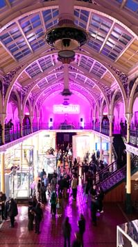 Lots of visitors in two level gallery which has an ironwork ceiling and balcony. The gallery is lit up in pink neon colours.