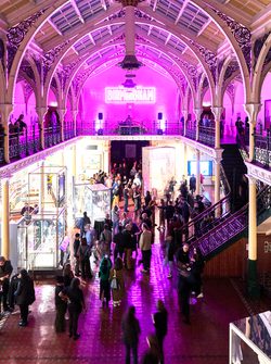 Lots of visitors in two level gallery which has an ironwork ceiling and balcony. The gallery is lit up in pink neon colours.