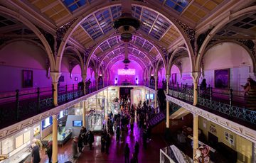 Lots of visitors in two level gallery which has an ironwork ceiling and balcony. The gallery is lit up in pink neon colours.