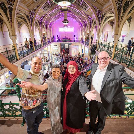 Four people standing with their arms out in a balcony with people in the gallery below.