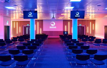 A large room set up in a theatre style seating arrangement, with projector, TV screens and podium. The room is lit with pink and blue lighting.