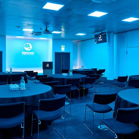A room set up in a cabaret seating arrangement with projector and podium. The room is lit with blue lighting.