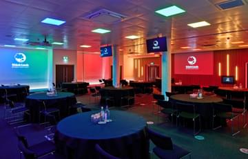Cabaret seating arrangement with two projectors, TV screens and podium. The room is lit with blue and red lighting.