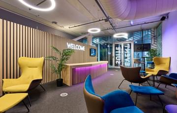 A modern reception area with reception desk, neon 'Welcome' sign and seating areas. The room is lit with purple lighting.