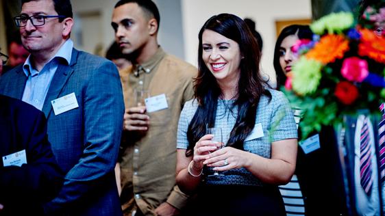 A group of people wearing name tags and holding drinks. A lady in the centre is smiling.