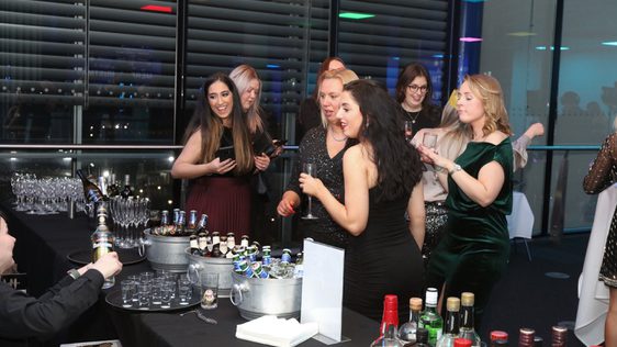 A group of people standing at a bar table full of drinks.
