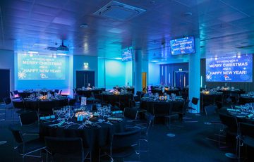 A wide shot of the events suite set up with circular tables which are laid out ready, the lighting is dark and blue.